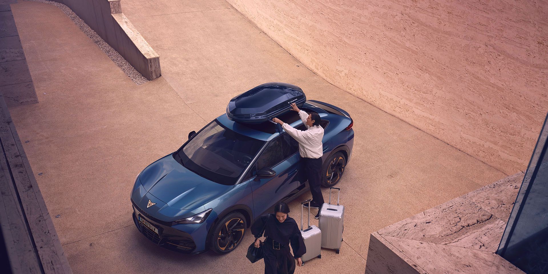 Eine Person greift zur Dachbox eines blauen CUPRA Tavascan. Zwei Koffer stehen neben dem Fahrzeug und eine Frau geht vom Fahrzeug weg.