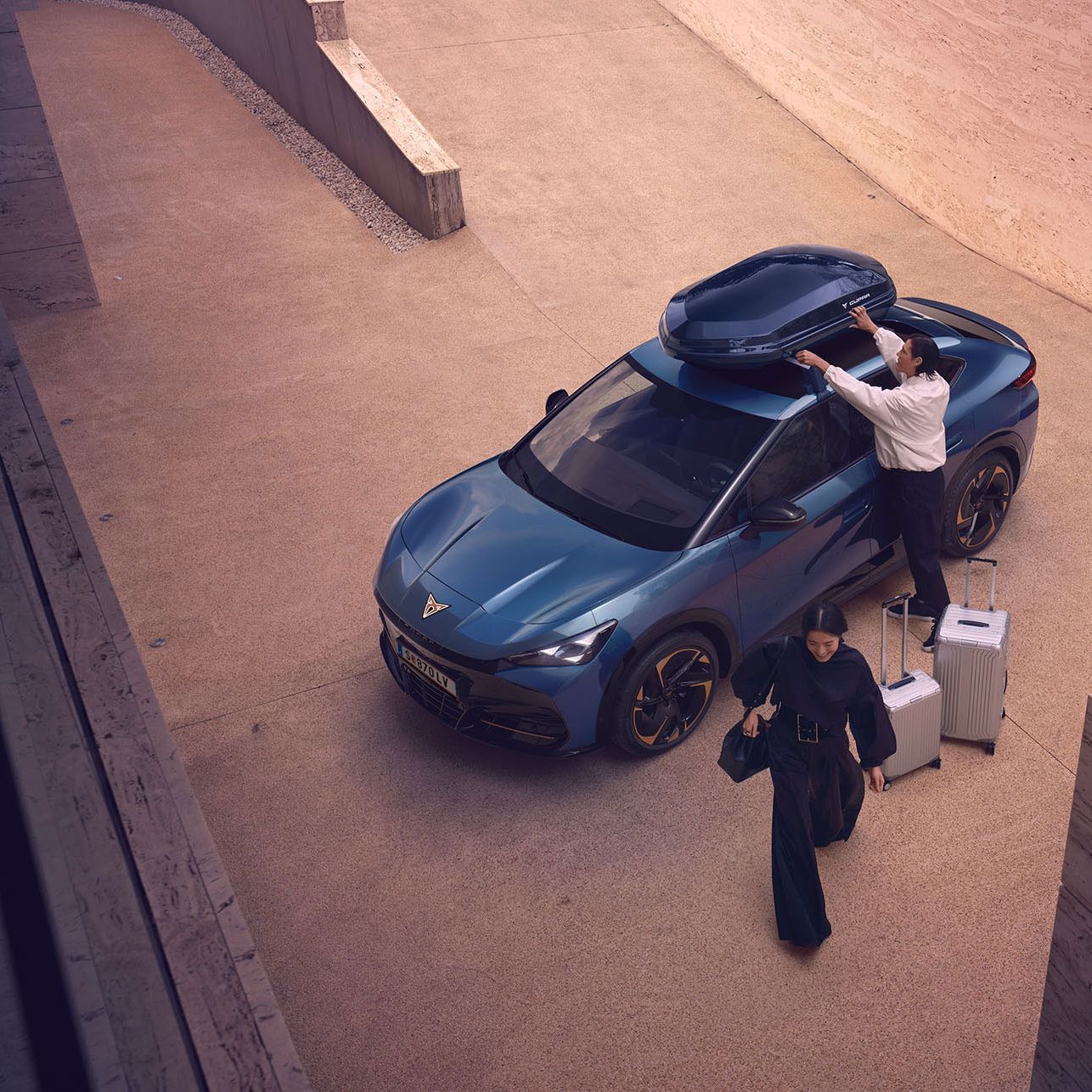 Eine Person greift zur Dachbox eines blauen CUPRA Tavascan. Zwei Koffer stehen neben dem Fahrzeug und eine Frau geht vom Fahrzeug weg.