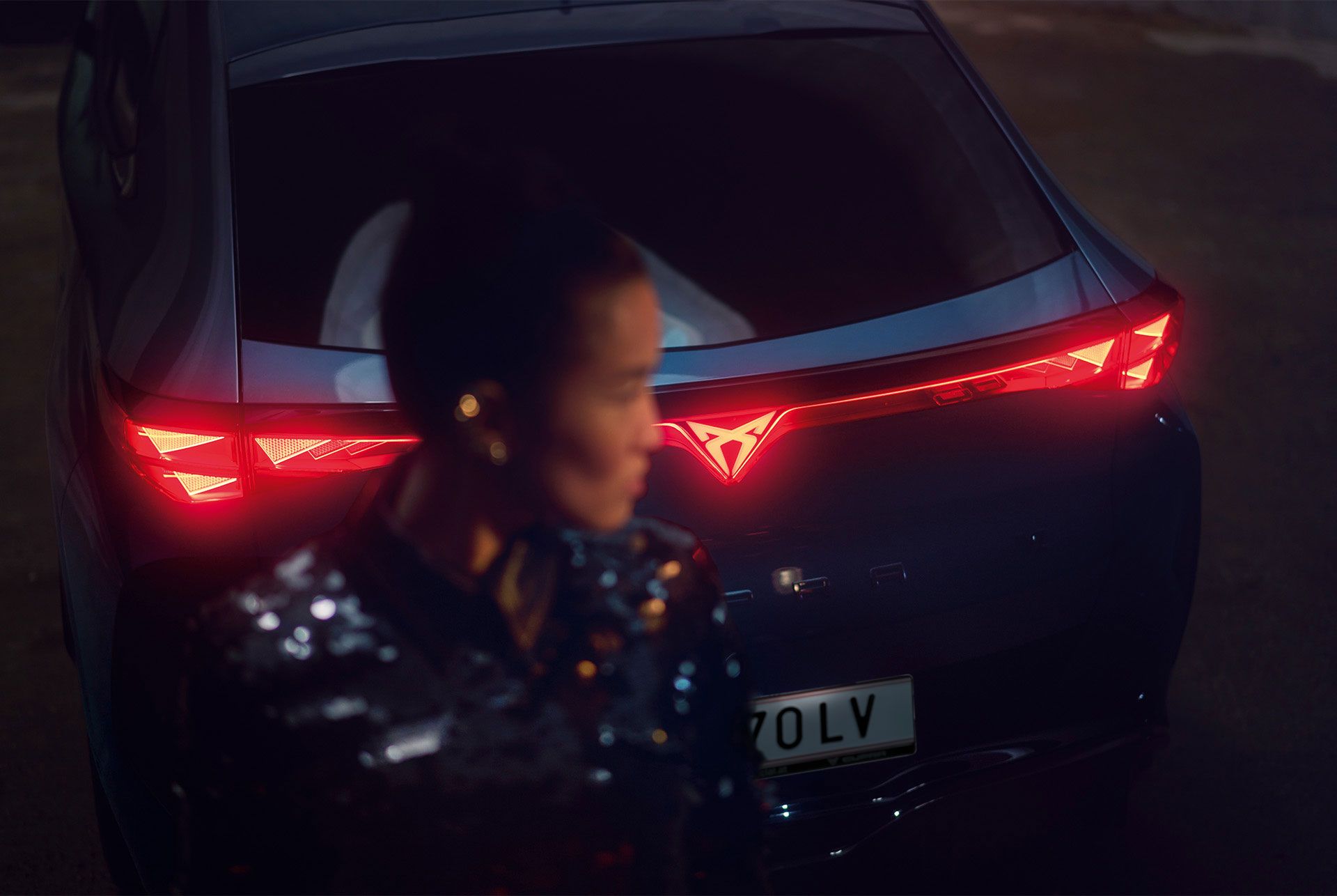Detailaufnahme vom Heck eines CUPRA Tavascan mit durchgehender Heckbeleuchtung, eine Frau steht neben dem Fahrzeug.