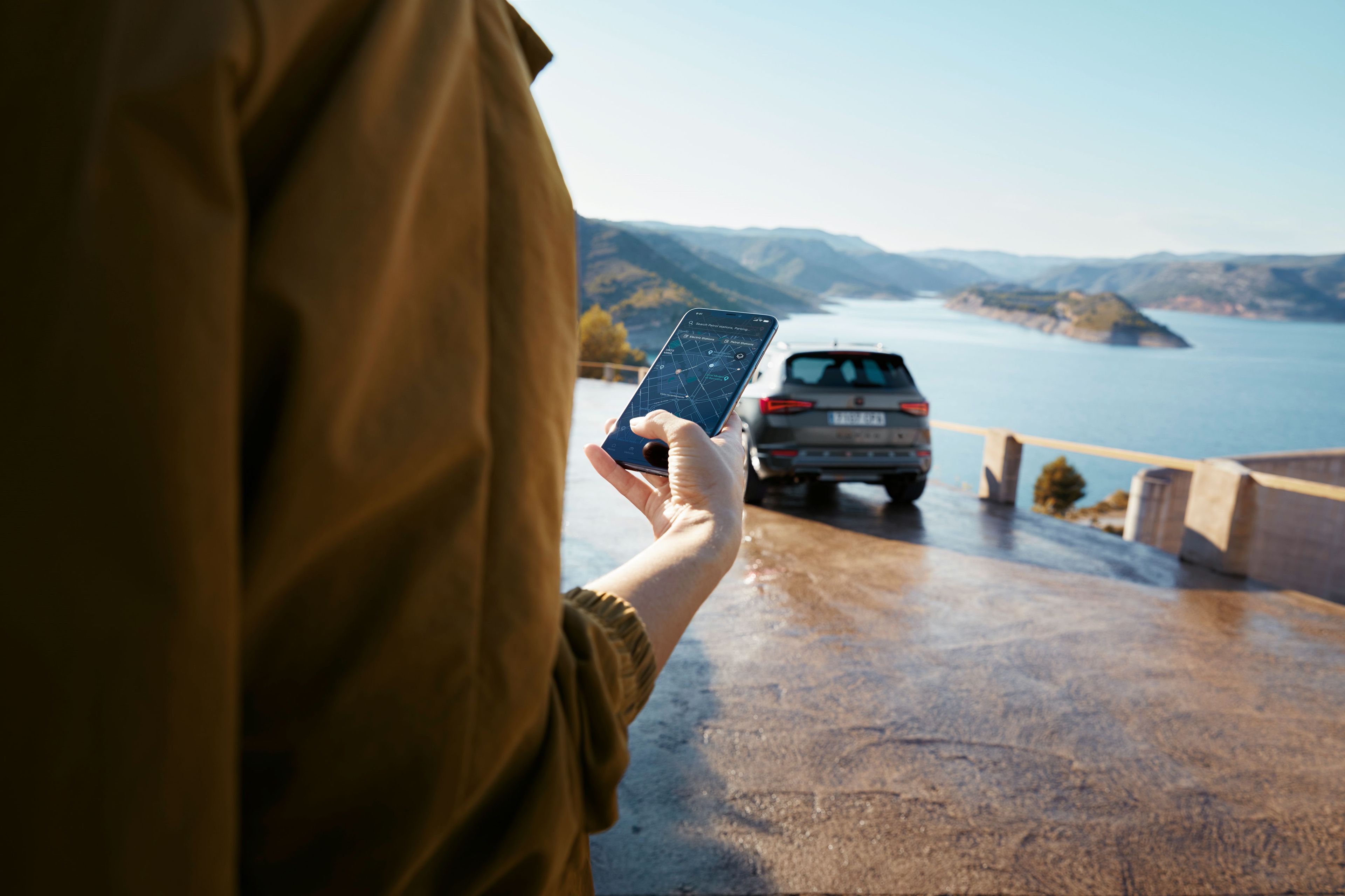 Eine Person hält ein Smartphone mit dem CUPRA Ateca im Hintergrund, der in der Nähe einer malerischen Aussicht auf Berge und Wasser parkt, was eine vernetzte Welt symbolisiert.